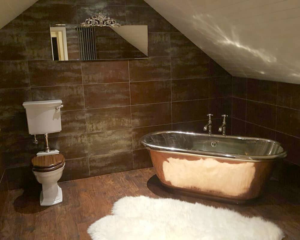 Copper freestanding bath with brown tiles, wooden floor and classic toilet and a white sheepskin rug